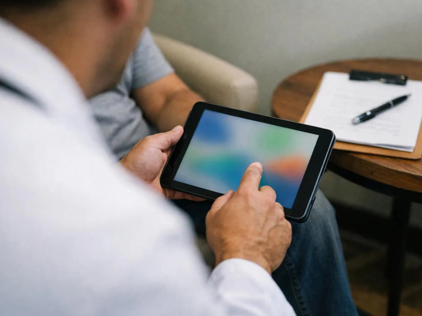 A person holds a tablet and taps the screen while sitting beside another person and a notepad on a small table