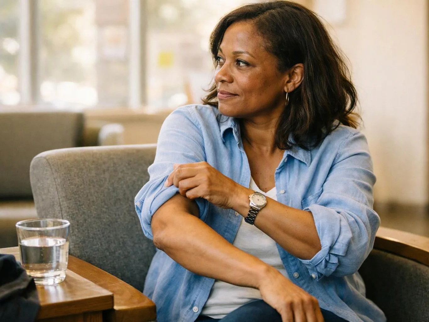 A woman in a blue shirt sits on a chair and rolls up her sleeve beside a glass of water